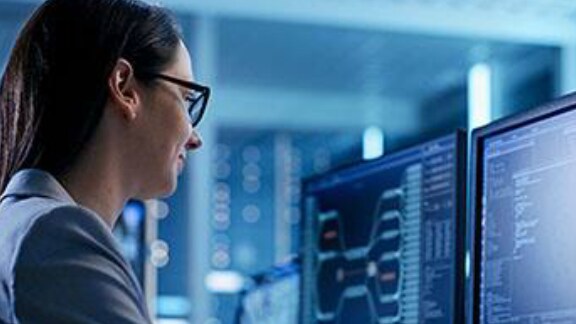 A woman wearing glasses, looking at two monitors displaying code