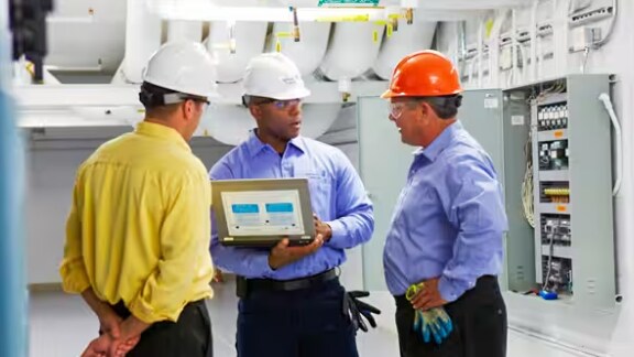 Three engineers in safety helmets discussing in a facility, while one of them holds a laptop
